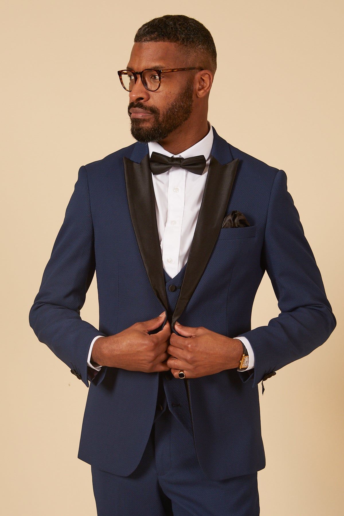 Charlie Wrenshall as Liam Connor wears the marcdarcy DALTON Royal Blue Tux Suit, a three-piece ensemble with black lapels, paired with a white shirt, black bowtie, and pocket square as he clasps his hands on his jacket.