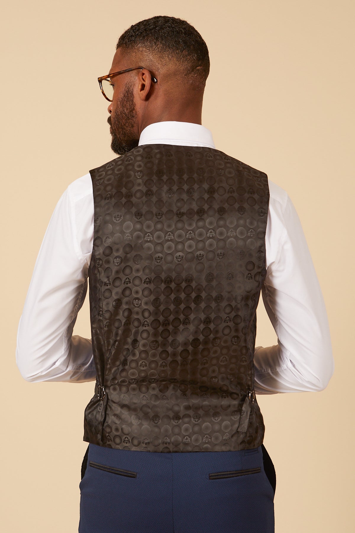 Back view of a man in a white dress shirt, marcdarcy DALTON Royal Blue Diamond Waistcoat, and blue suit pants. He has short dark hair, a beard, and wears tortoise-shell glasses.