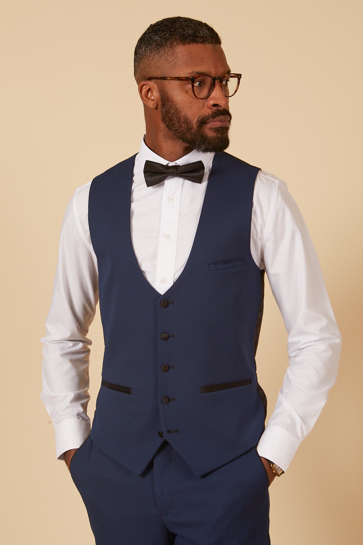 A man wearing the marcdarcy DALTON Royal Blue Diamond Waistcoat, paired with a white shirt and black bow tie, looks to the right. He has glasses and a beard.