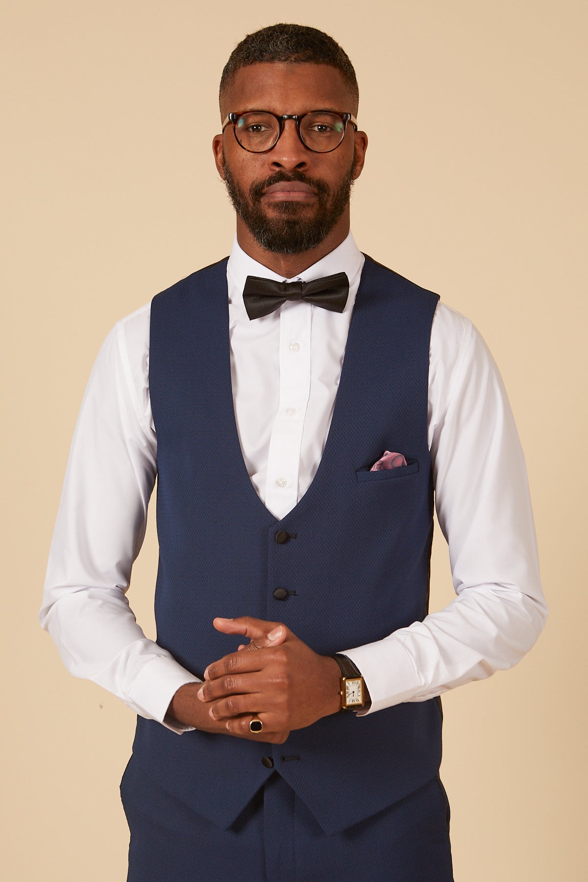 A man with short hair and a beard wears glasses, a white dress shirt, the marcdarcy DALTON Royal Blue Diamond Waistcoat, a black bow tie, watch, and ring while posing against a plain beige background.