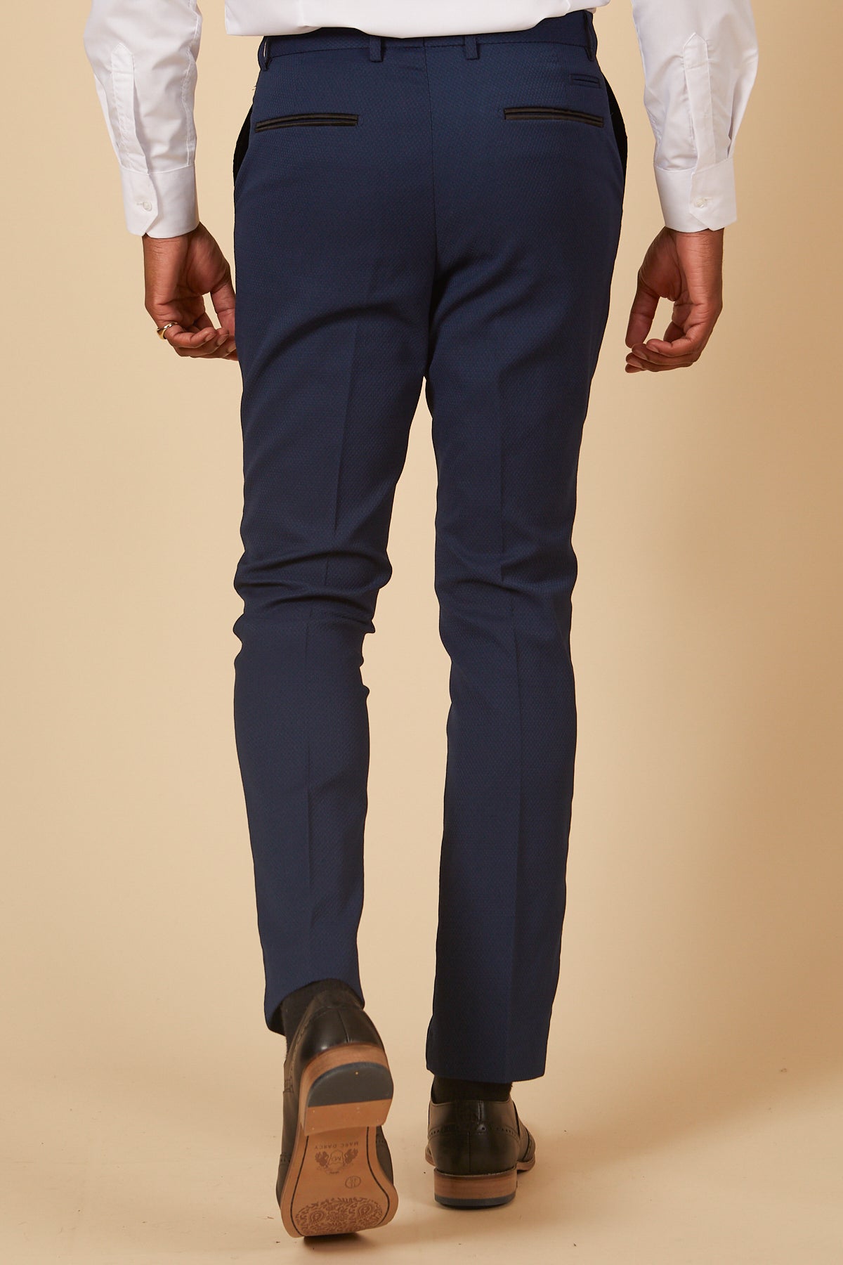 A man in formal attire walks away in brown dress shoes, wearing a white shirt and marcdarcy’s DALTON - Royal Blue Diamond Trousers. The beige background enhances the polished look.