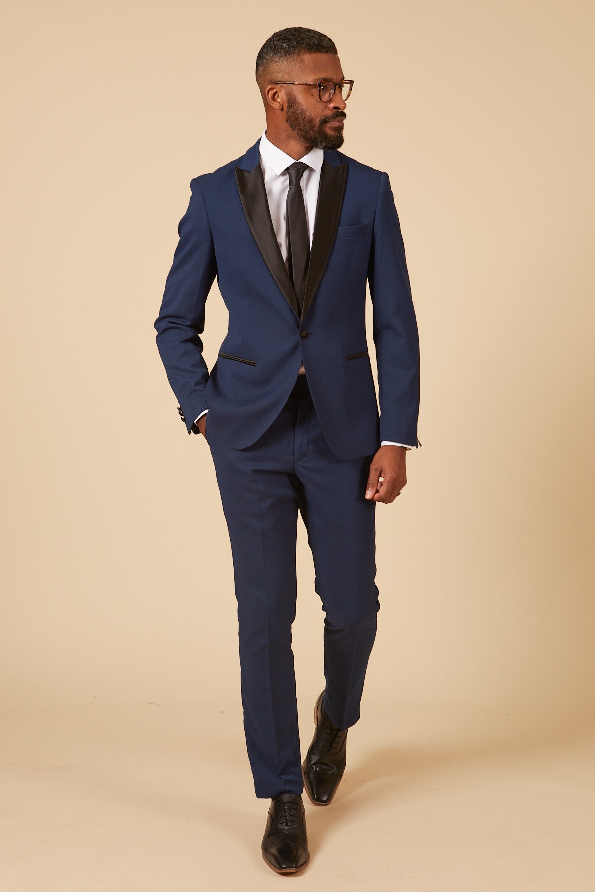A Black man in a slim fit marcdarcy DALTON Royal Blue Tux Lapel Diamond Blazer with a satin lapel and black tie stands with his hand in his pocket, looking to the right.