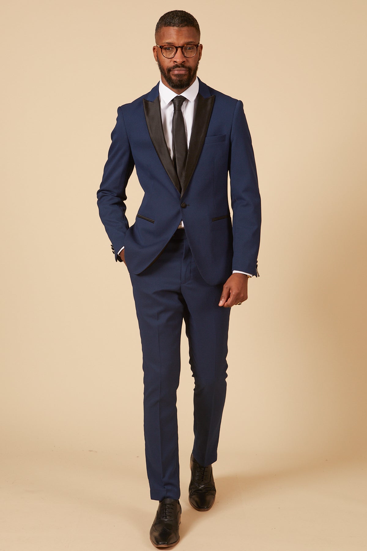 A man in the marcdarcy DALTON - Royal Blue Tux Lapel Diamond Blazer, white shirt, black tie, and black shoes walks confidently toward the camera against a beige background—ideal for formal occasions.