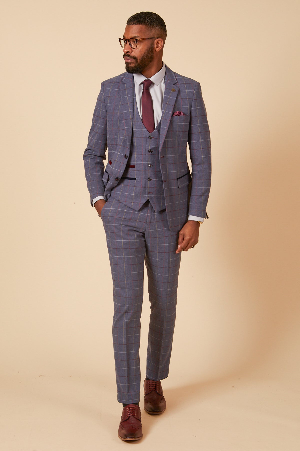 A man in the marcdarcy DRAKE Sky Blue Check Three Piece Suit, paired with a maroon tie and brown dress shoes, stands against a beige background, looking to the side with one hand in his pocket—ideal formalwear.
