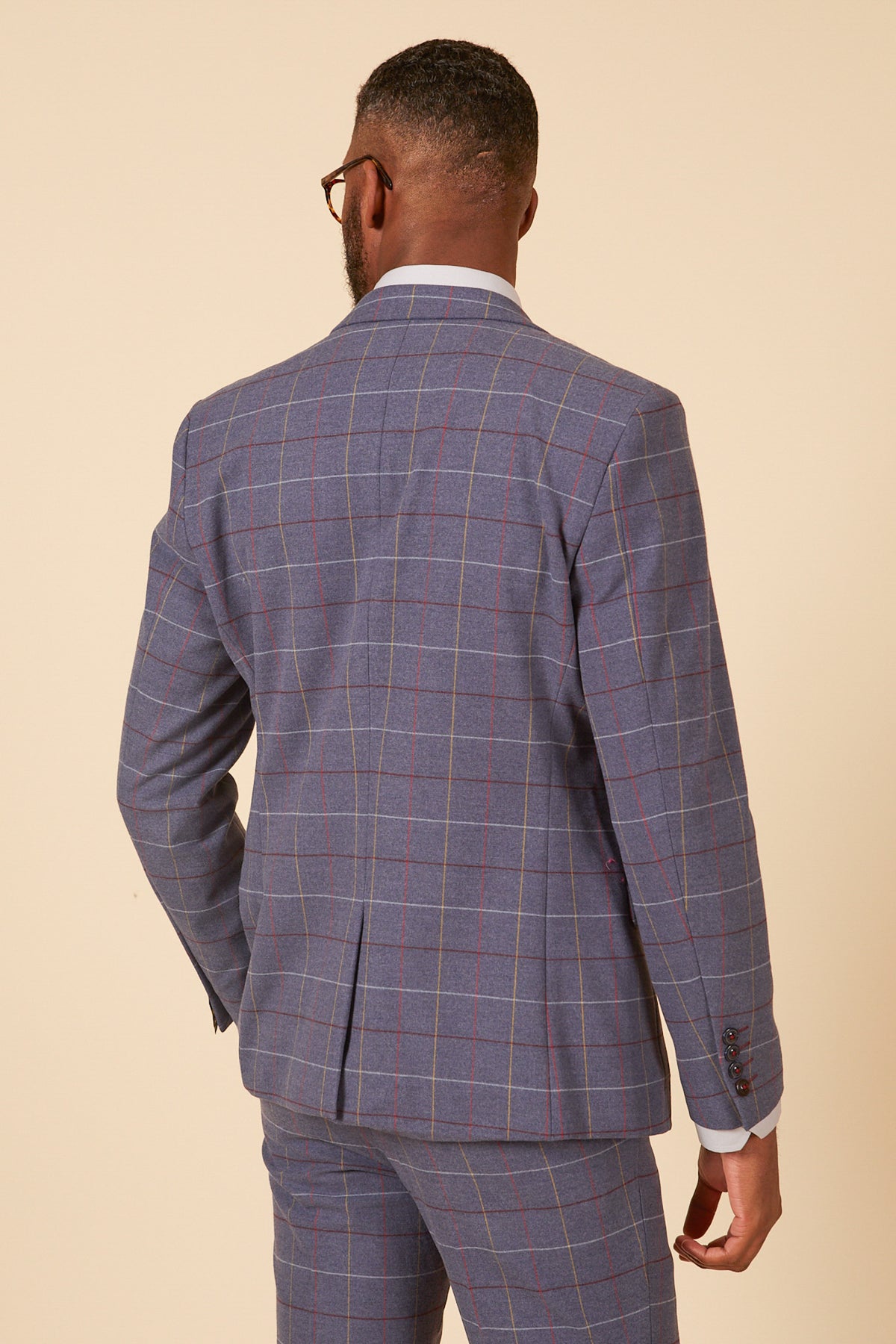 A man with short hair and glasses stands with his back to the camera in a marcdarcy DRAKE Sky Blue Check Three Piece Suit, highlighting the suit’s detail against a plain, neutral background.