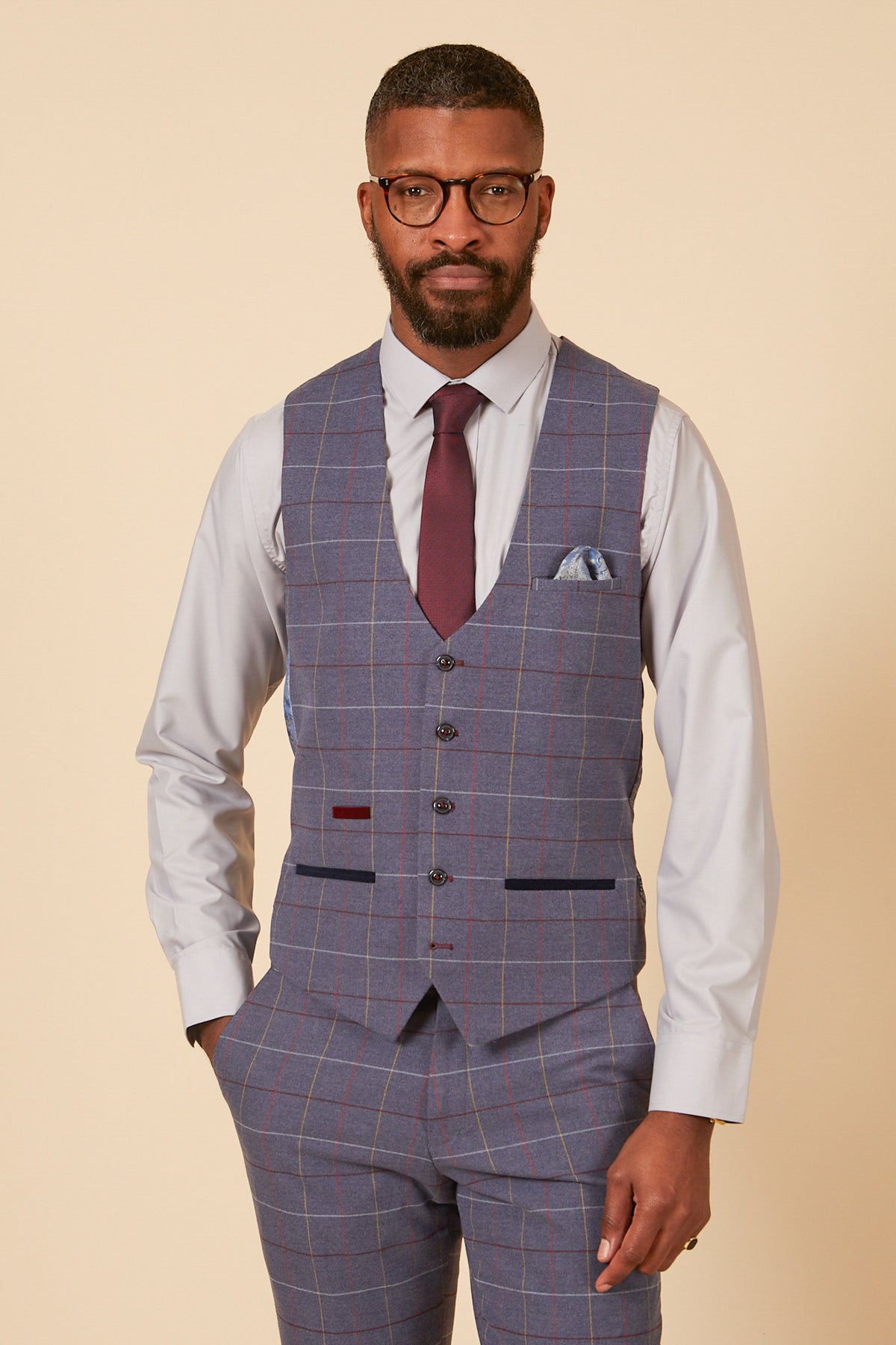 A man in the marcdarcy DRAKE - Sky Blue Check Three Piece Suit—featuring a light blue shirt, check jacket, matching vest and pants—poses against a beige background, finishing his formal look with a maroon tie and pocket square.