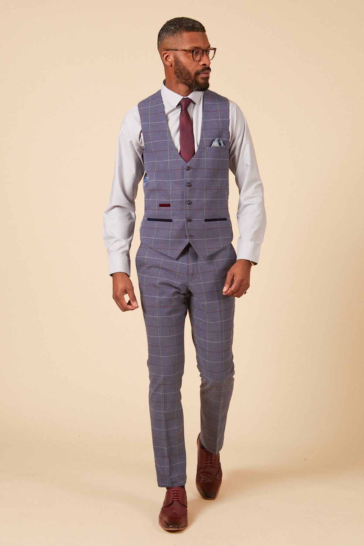 A man models the marcdarcy DRAKE - Sky Blue Check Three Piece Suit with a blue plaid vest, matching pants, burgundy tie, pocket square, and brown shoes against a beige background—perfect for any formal event.