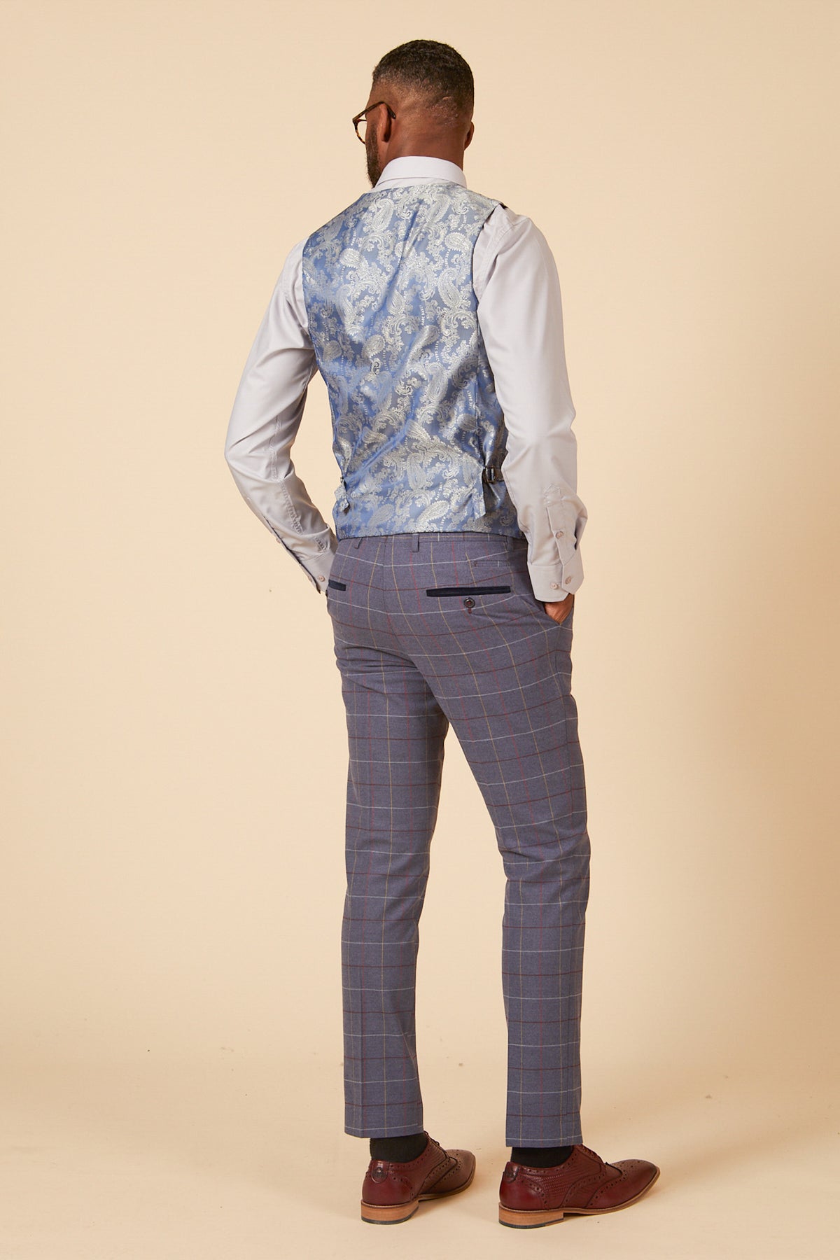 A man wears the marcdarcy DRAKE - Sky Blue Check Three Piece Suit, featuring a sky blue checked vest over a light blue shirt and plaid pants, paired with brown dress shoes—a refined choice for any formal event.