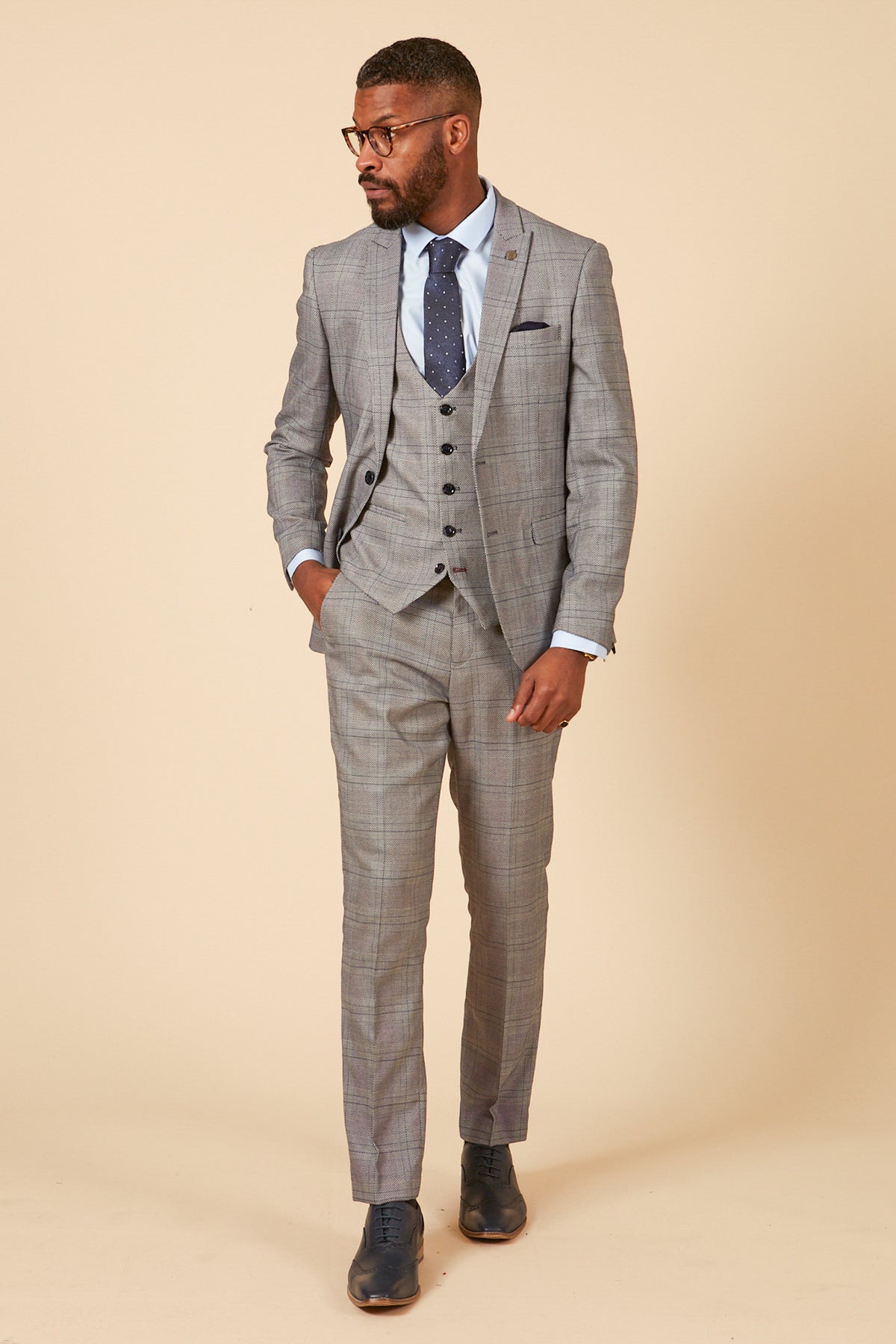 A man wearing the marcdarcy JERRY Grey Check Suit With Single Breasted Waistcoat, paired with a white shirt, navy polka dot tie, and dark shoes stands against a beige background with one hand in his pocket.
