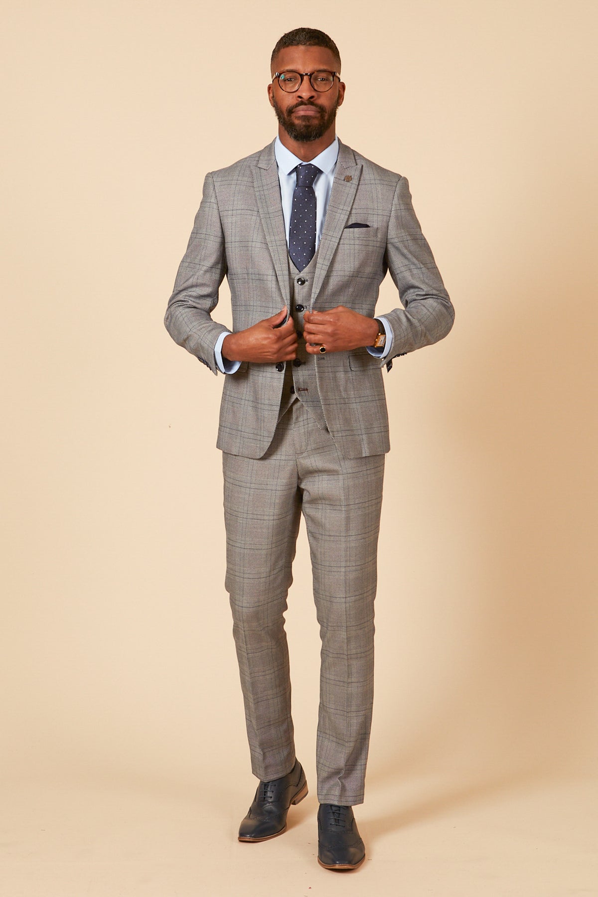 A man in the marcdarcy JERRY Grey Check Suit with a single-breasted waistcoat, blue dress shirt, navy tie, and navy dress shoes stands buttoning his jacket against a beige background.