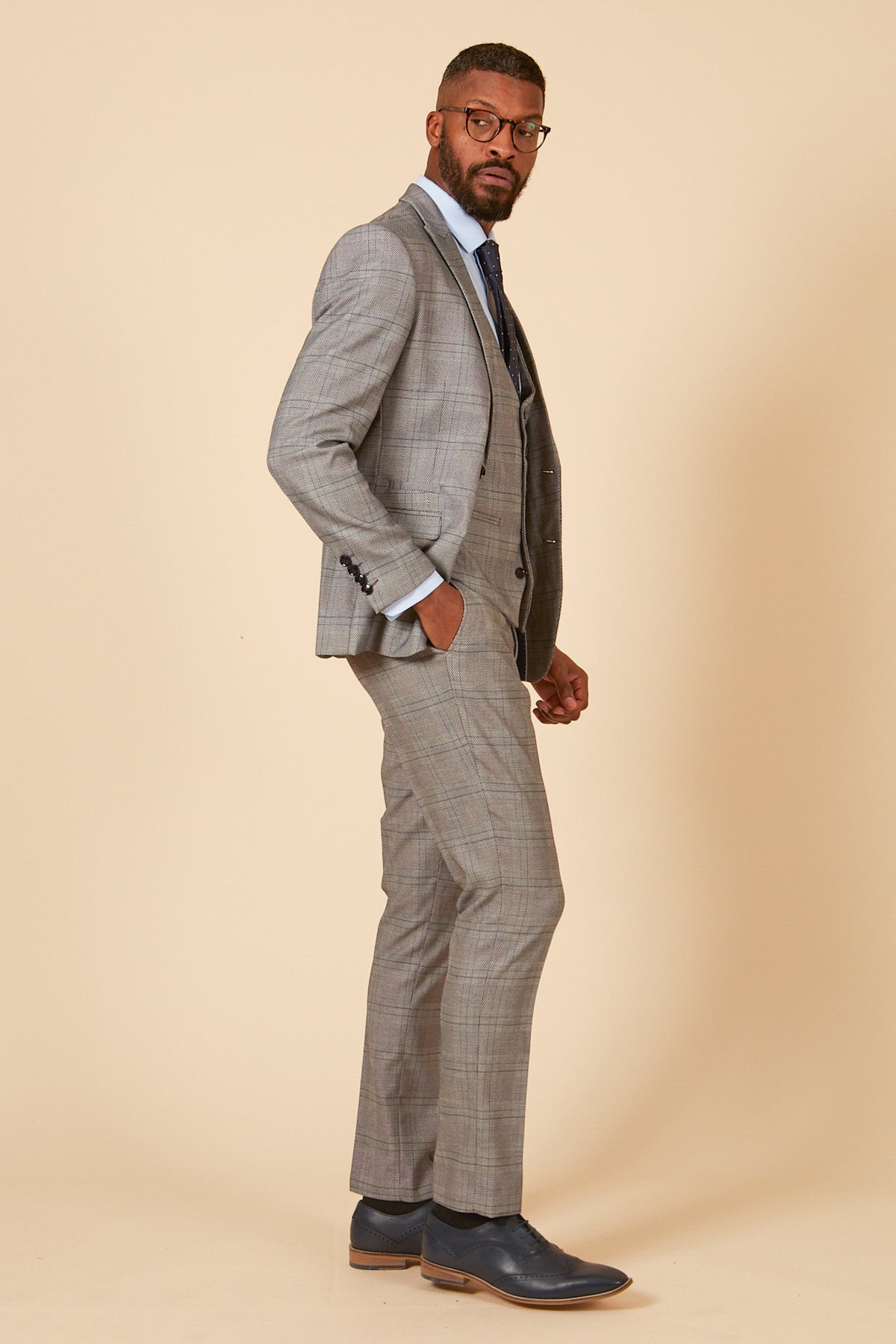A man wearing the marcdarcy JERRY Grey Check Suit with Single Breasted Waistcoat stands sideways with one hand in his pocket, looking at the camera against a beige background.