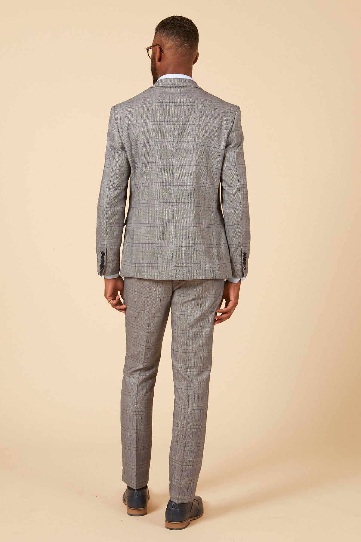 A man models the marcdarcy JERRY Grey Check Suit with Single Breasted Waistcoat, paired with a white shirt and dark shoes, facing away from the camera against a plain beige background.