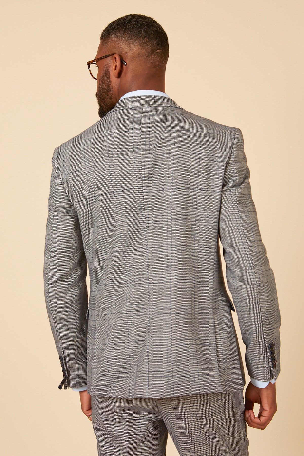 A man with short hair, a trimmed beard, and glasses faces away in a marcdarcy JERRY Grey Check Blazer. The slim fit blazer features a peak lapel and plaid pattern, set against a plain light beige background.