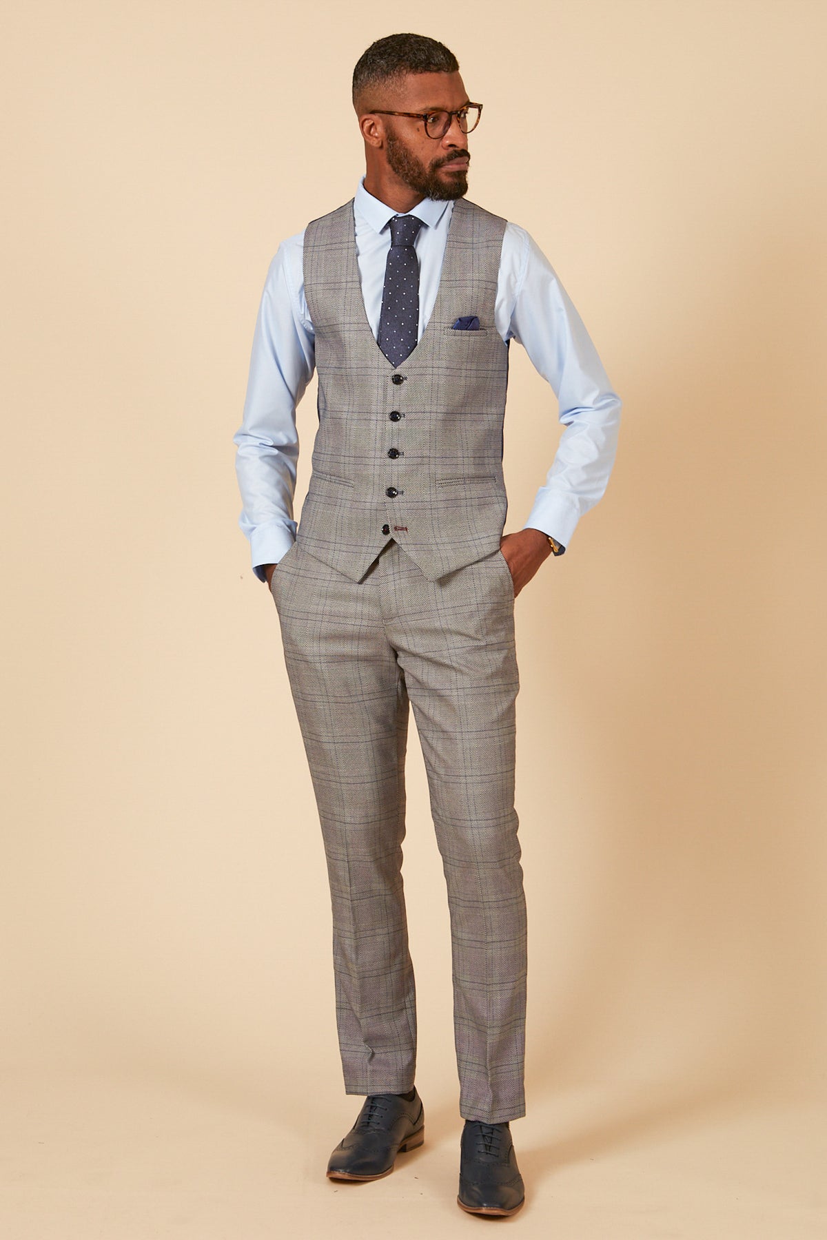 A man stands with hands in pockets, wearing the marcdarcy JERRY Grey Check Suit with a single breasted waistcoat, light blue shirt, navy polka dot tie, glasses, and dark dress shoes against a beige background.