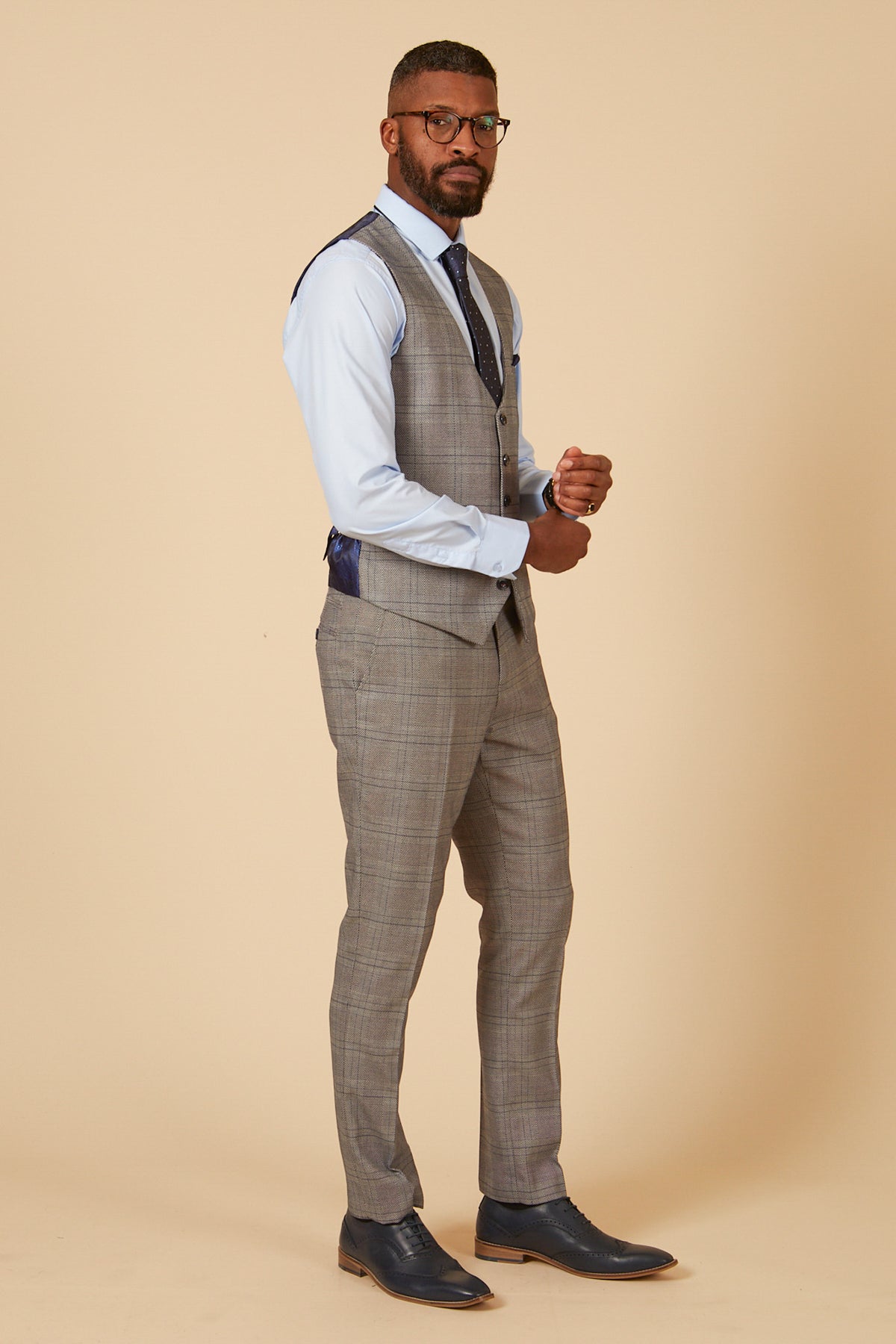 A man wearing glasses stands against a beige background in a light blue dress shirt, grey check JERRY single breasted waistcoat by marcdarcy, matching trousers, navy tie, and navy dress shoes.