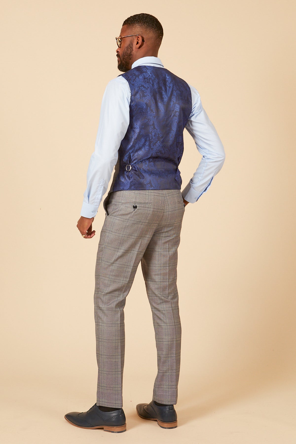 A man faces away, wearing the marcdarcy JERRY grey check suit with a single breasted waistcoat, paired with a light blue shirt and dark shoes, set against a beige background.