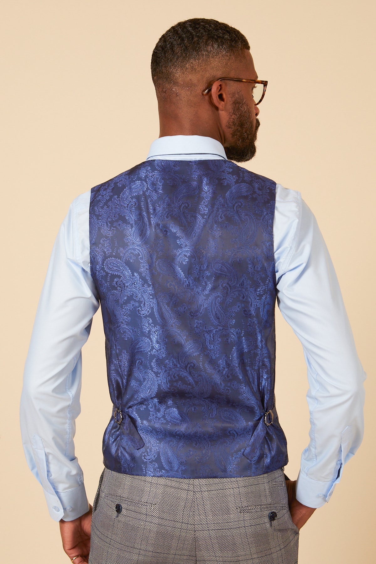 A man wearing glasses, a light blue dress shirt, and the marcdarcy JERRY - Grey Check Single Breasted Waistcoat stands facing away from the camera against a plain beige background.