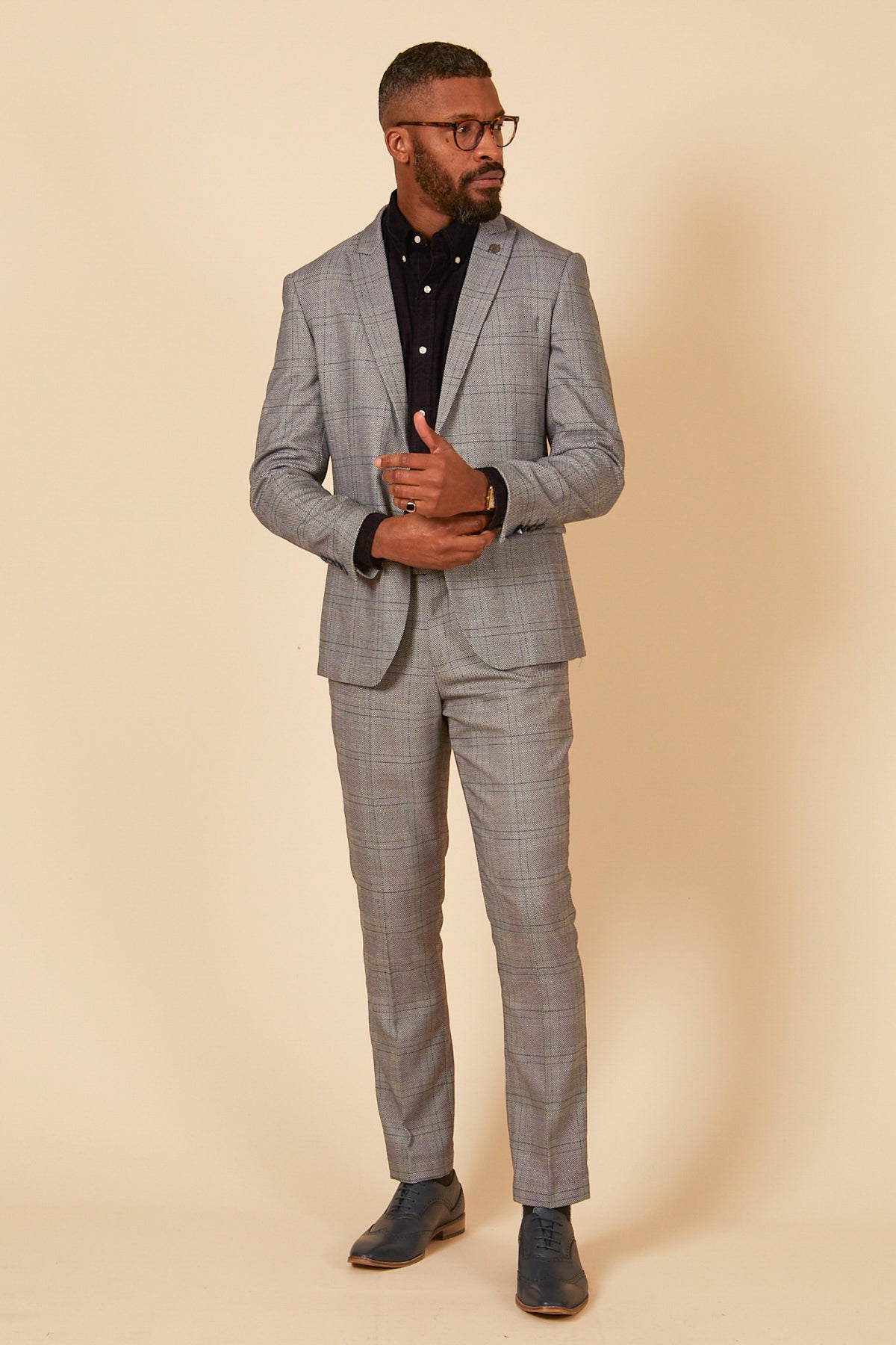 A man wearing glasses stands against a beige background in the marcdarcy JERRY - Grey Check Blazer, a slim fit suit with peak lapels, paired with a black button-up shirt and dark shoes, looking slightly left with his hands clasped.