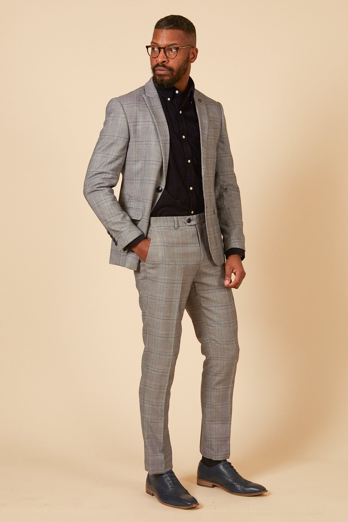 A man with short hair, a beard, and glasses stands against a beige background wearing the marcdarcy JERRY - Grey Check Blazer, paired with a black button-up shirt and dark dress shoes, one hand in his pocket.