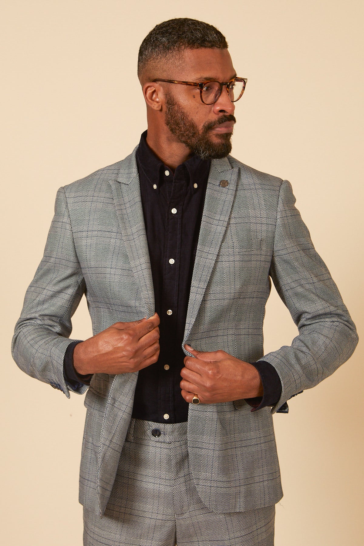 A man with short hair, a beard, and glasses wears the marcdarcy JERRY Grey Check Blazer over a black button-up shirt. He adjusts his jacket while looking to the side against a plain beige background.