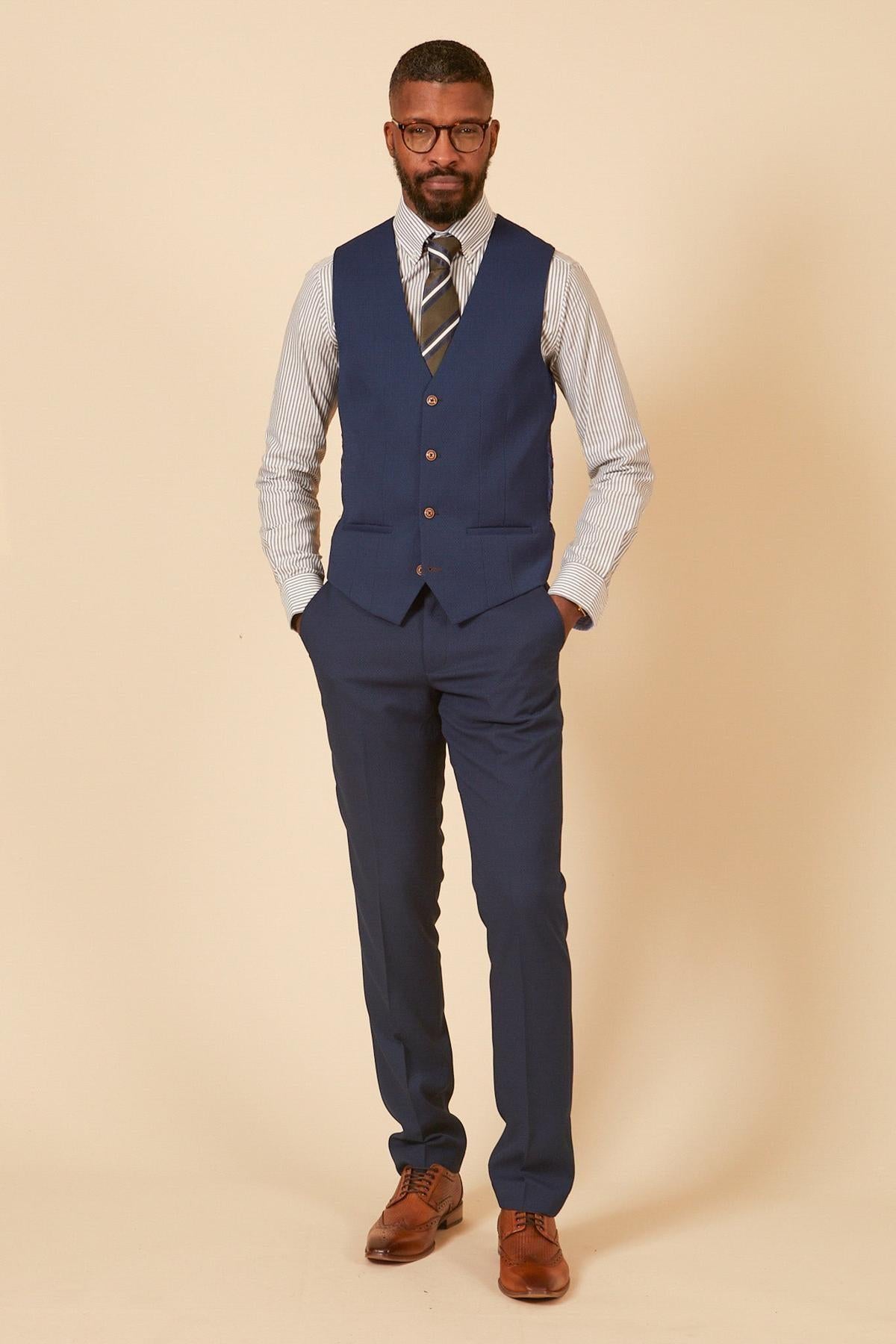 A man in a marcdarcy MAX Royal Blue Single Breasted Waistcoat, striped shirt, tie, and brown dress shoes stands against a beige background with hands in pockets—ideal office wear. He has short hair, a beard, and glasses.