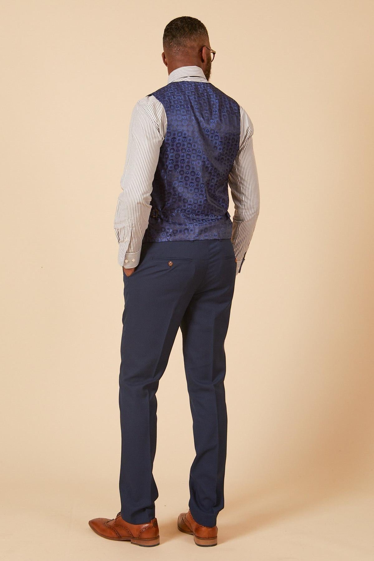 A man stands facing away, wearing a marcdarcy MAX Royal Blue Single Breasted Waistcoat over a striped shirt, blue dress pants, and brown dress shoes against a beige background.