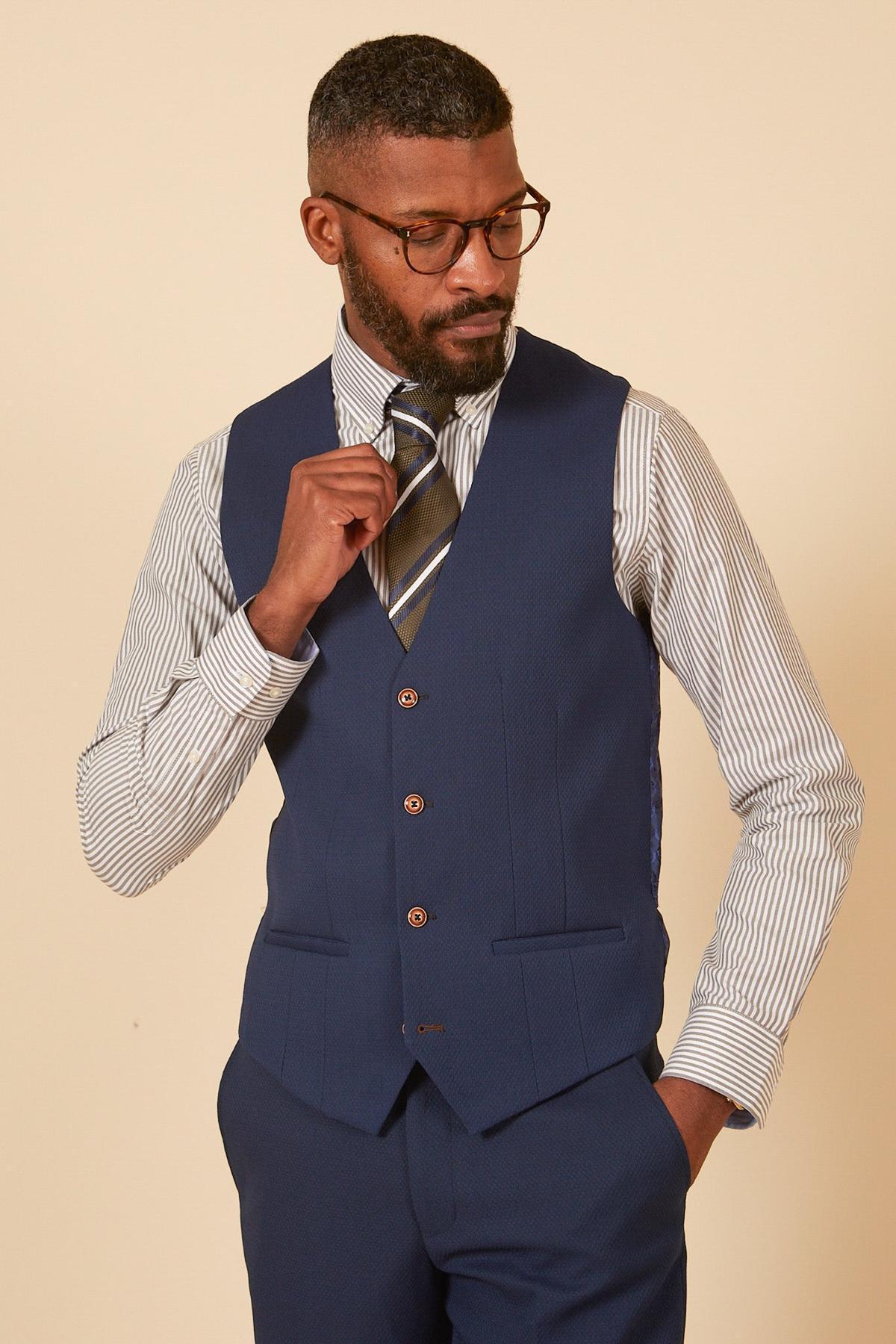 A man in glasses wears a marcdarcy MAX Royal Blue Single Breasted Waistcoat with matching trousers, a white shirt with black stripes, and a striped tie, adjusting his tie while looking down to his left—ideal office attire.