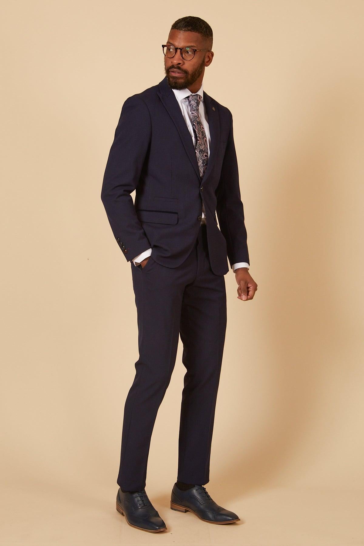 A man wearing the Marc Darcy Cardiff City F.C. Collection - BROMLEY Navy Check Two Piece Suit as worn by Will Fish, a white dress shirt, patterned tie, and dress shoes stands with one hand in his pocket against a beige background.
