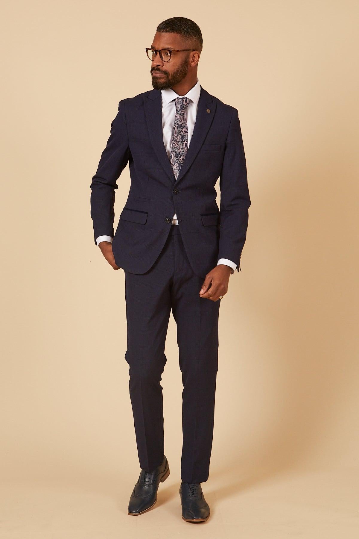 A man in the Marc Darcy BROMLEY Navy Check Two Piece Suit from The Cardiff City F.C. Collection, as worn by Will Fish, pairs it with a white shirt and paisley tie. He stands against a beige background, sporting glasses and a beard.