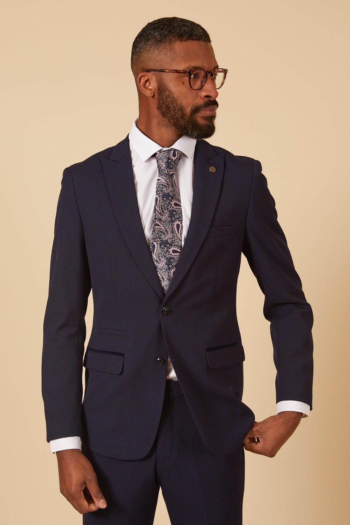 A man in the Marc Darcy Cardiff City F.C. Collection BROMLEY Navy Check Two Piece Suit, as worn by Will Fish, paired with a white shirt and patterned tie, stands against a beige background looking to the side. He has short hair, a beard, and glasses.