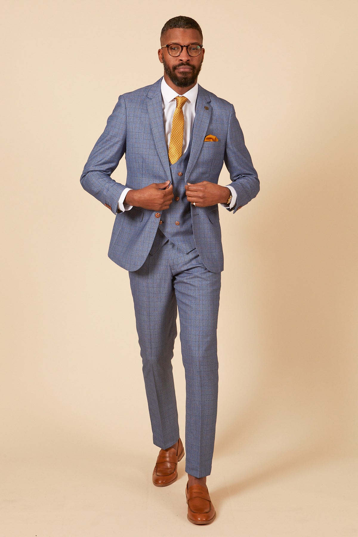 A man in the marcdarcy GEORGE Light Blue Check Three Piece Suit adjusts his jacket against a cream background, wearing a white shirt, yellow patterned tie, and brown shoes.