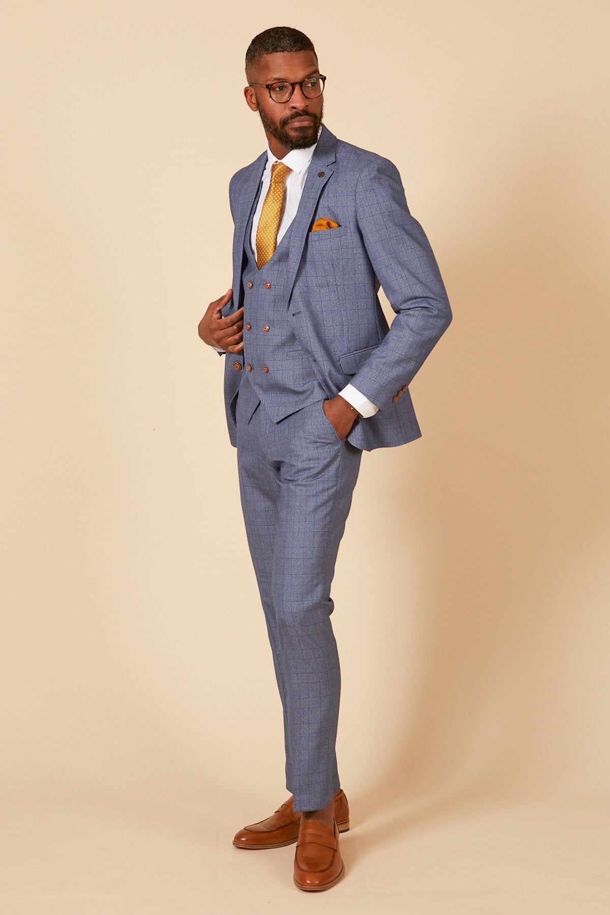 A man in the marcdarcy GEORGE Light Blue Check Three Piece Suit with a white shirt, yellow tie, pocket square, brown dress shoes, and glasses stands confidently against a beige background.