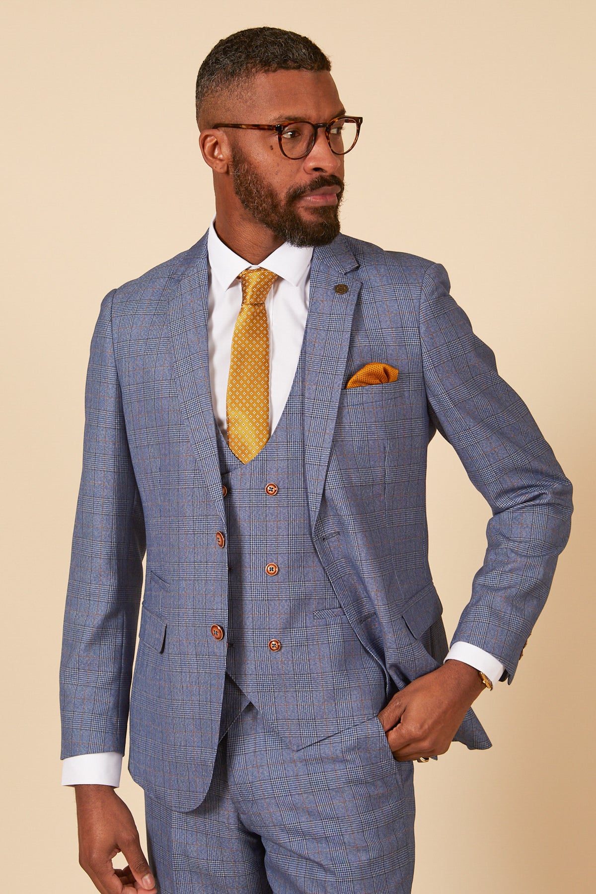 A man in the marcdarcy GEORGE Light Blue Check Three Piece Suit, white shirt, yellow patterned tie, and yellow pocket square stands confidently against a beige background. He has glasses and a trimmed beard.