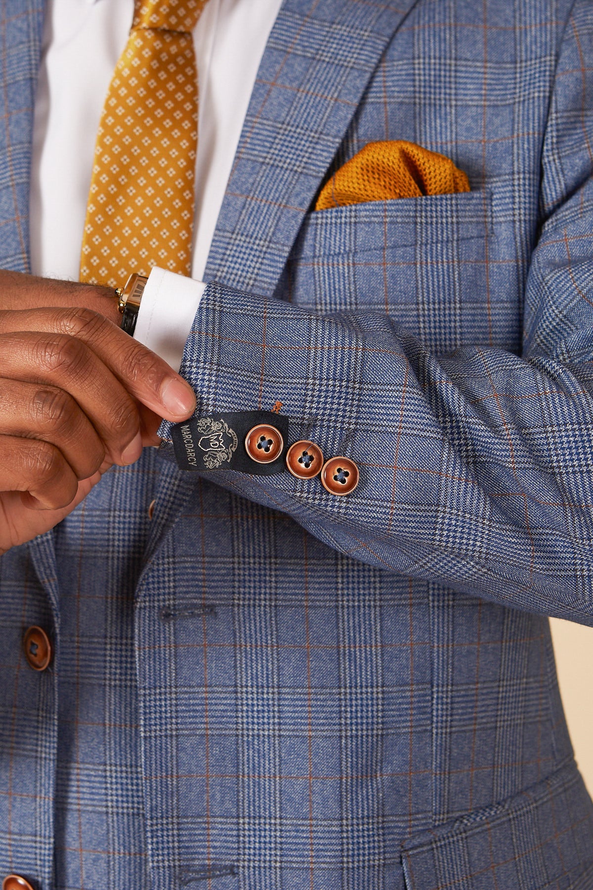 Close-up of a man's hand adjusting a marcdarcy GEORGE light blue check three piece suit, revealing a gold patterned tie and pocket square. The suit features brown buttons and the designer's signature label.