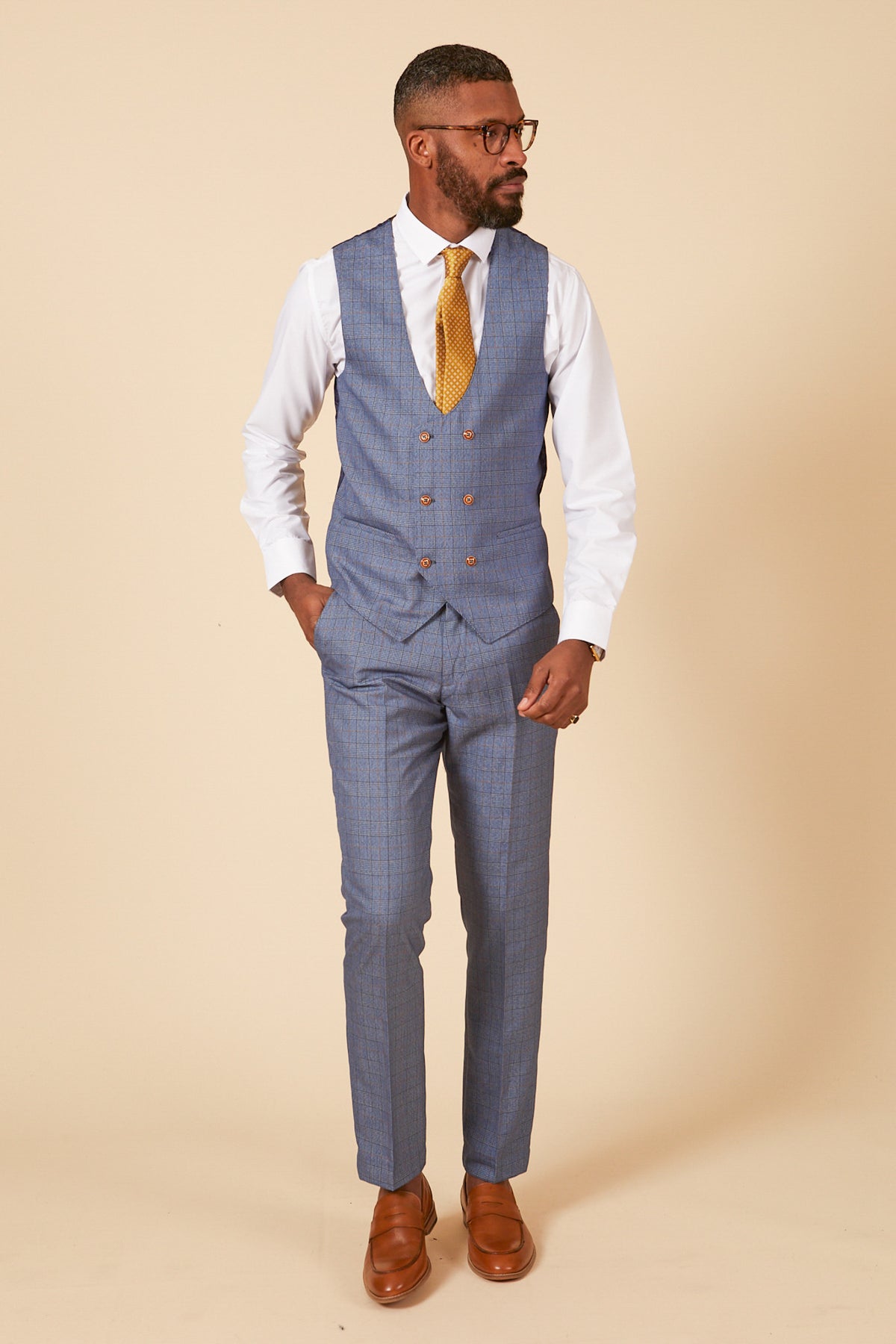 A man in glasses, a white shirt, yellow tie, and marcdarcy's GEORGE Light Blue Check Three Piece Suit with brown dress shoes stands against a beige background, one hand in his pocket.