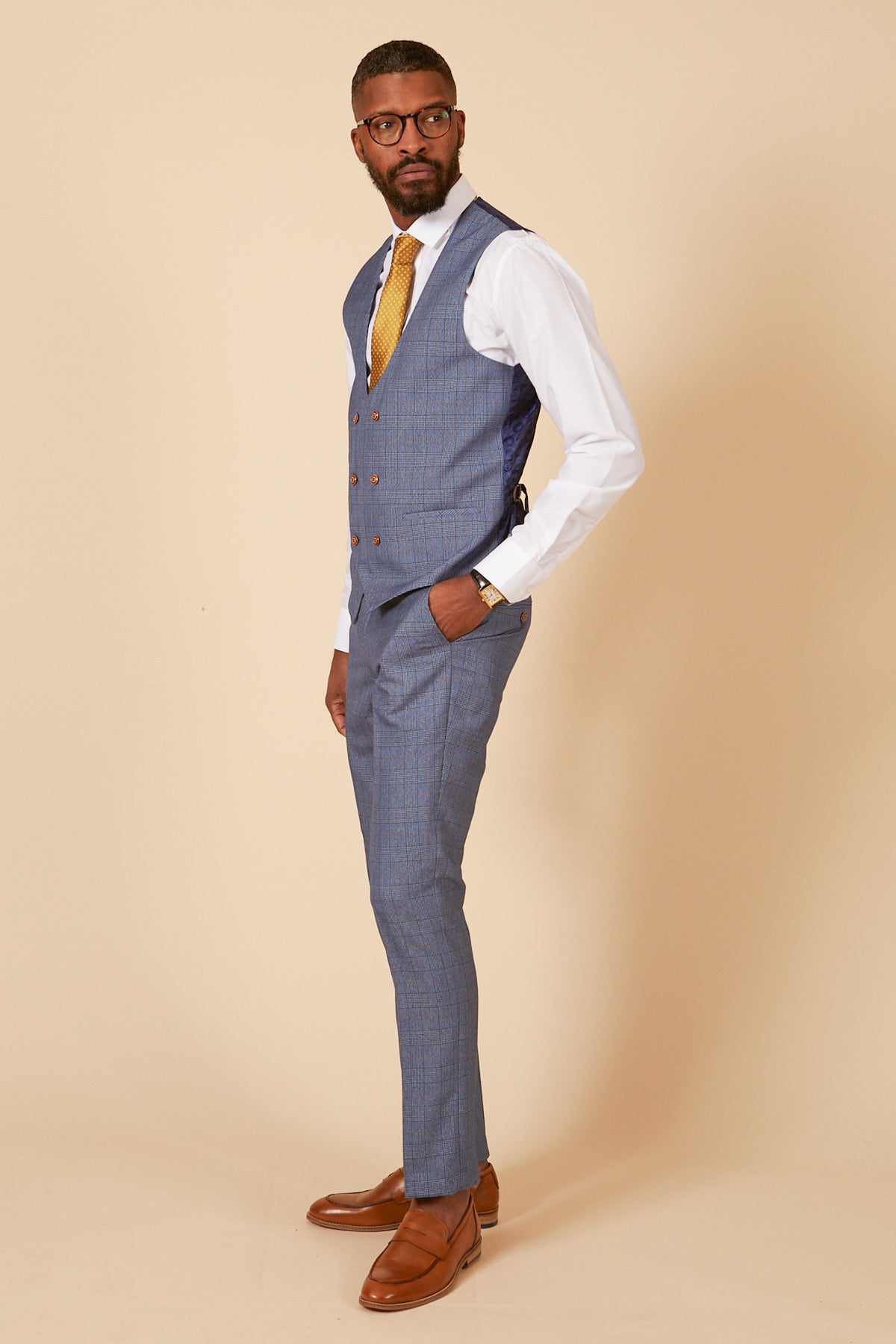 A man in glasses poses sideways against a beige background, wearing a marcdarcy LEE GEORGE light blue double-breasted waistcoat, blue checked suit, white shirt, yellow tie, and brown dress shoes.