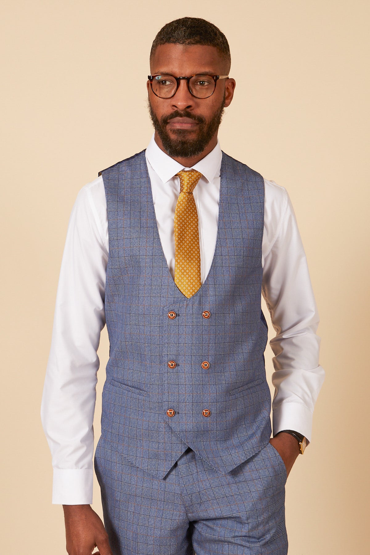 A man with short hair, a beard, and glasses wears the marcdarcy GEORGE light blue check three piece suit, styled with a white shirt and yellow tie, standing against a plain beige background.