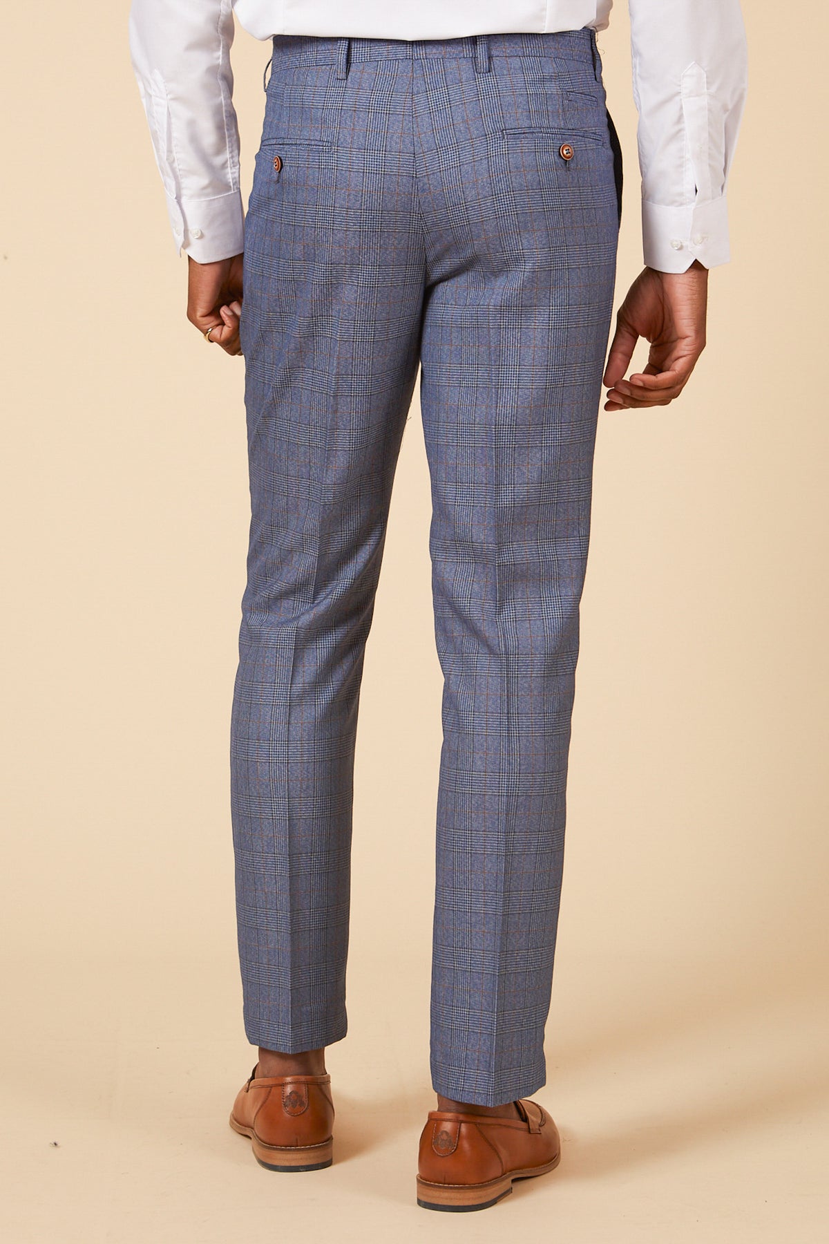 A man wearing the marcdarcy GEORGE Light Blue Check Three Piece Suit, with a white dress shirt and brown dress shoes, stands facing away from the camera against a beige background.