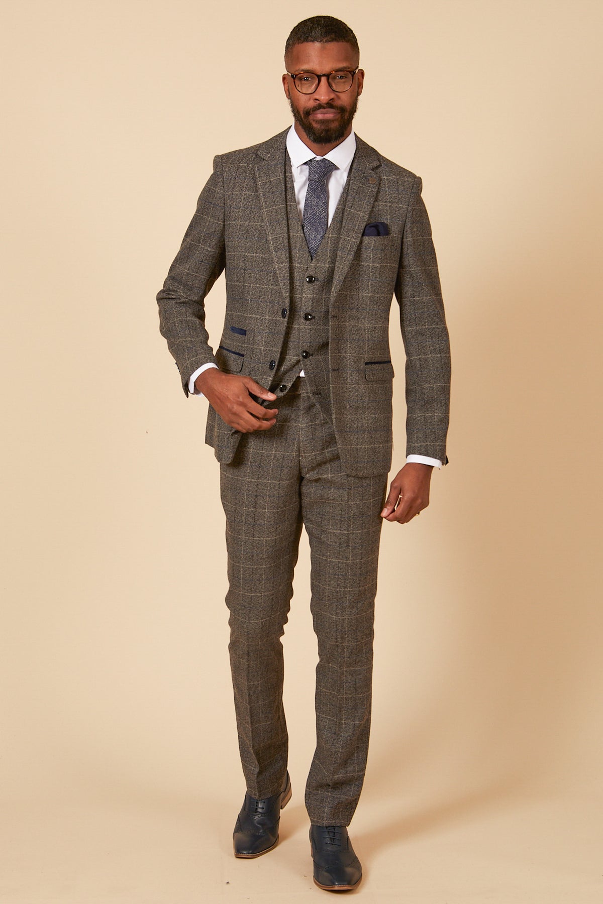 A man in the marcdarcy SCOTT Grey Tweed Check Three Piece Suit stands against a beige background, wearing glasses, a white shirt, blue tie, and dark shoes, looking confidently at the camera.