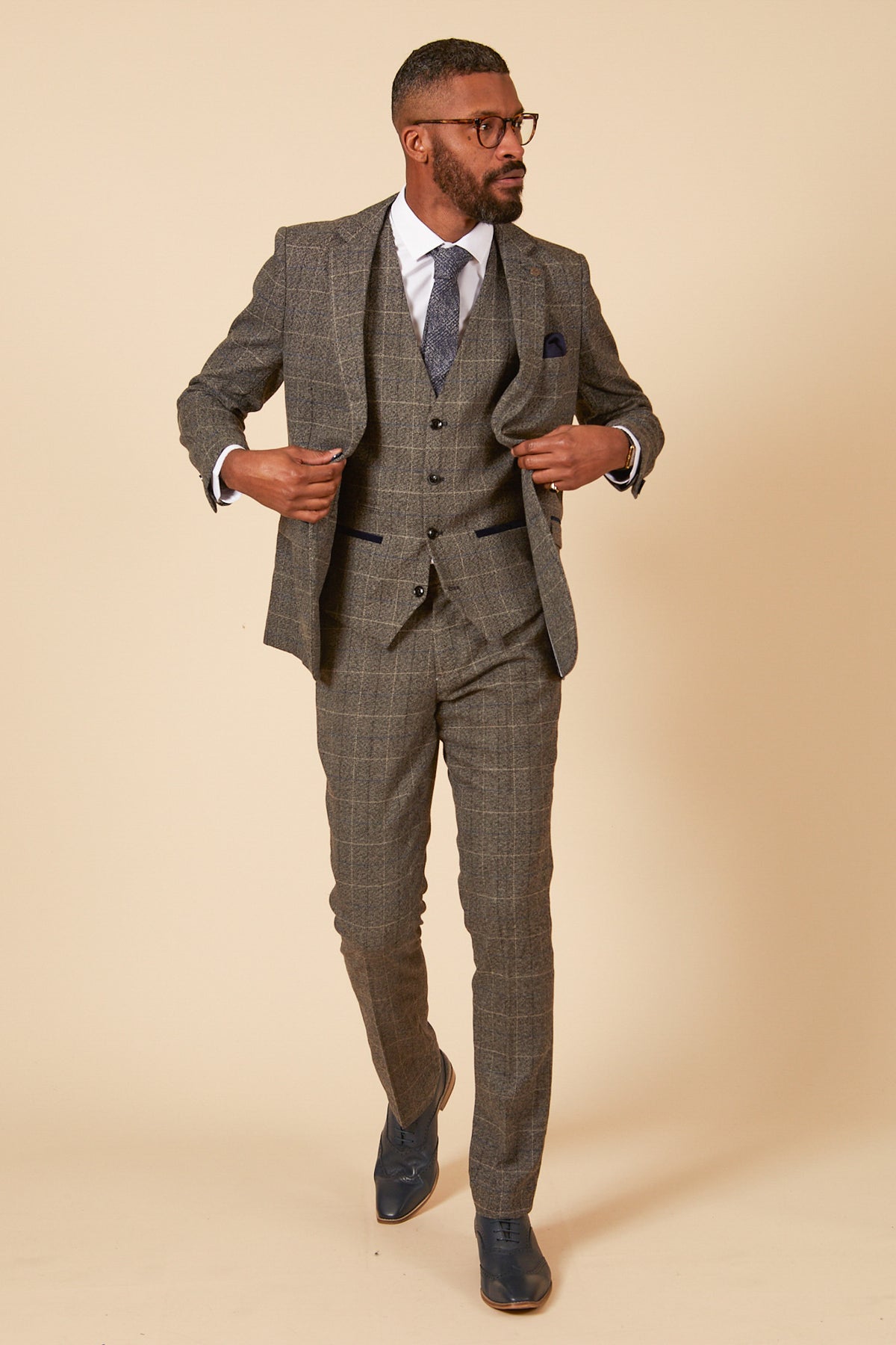 A man in glasses poses against a beige background wearing the marcdarcy SCOTT Grey Tweed Check Three Piece Suit with a white shirt, patterned tie, navy velvet pocket trims, and blue shoes.