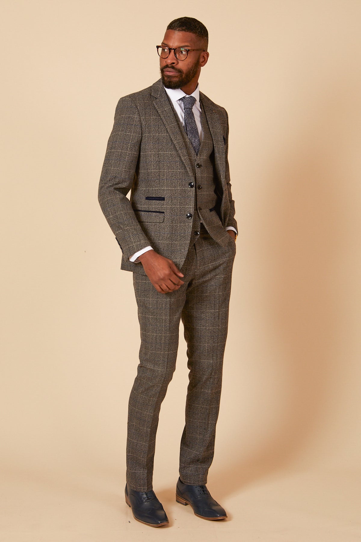 A man in the marcdarcy SCOTT Grey Tweed Check Three Piece Suit, styled with a white shirt and patterned tie, stands against a beige background, gazing to the side with one hand in his pocket.