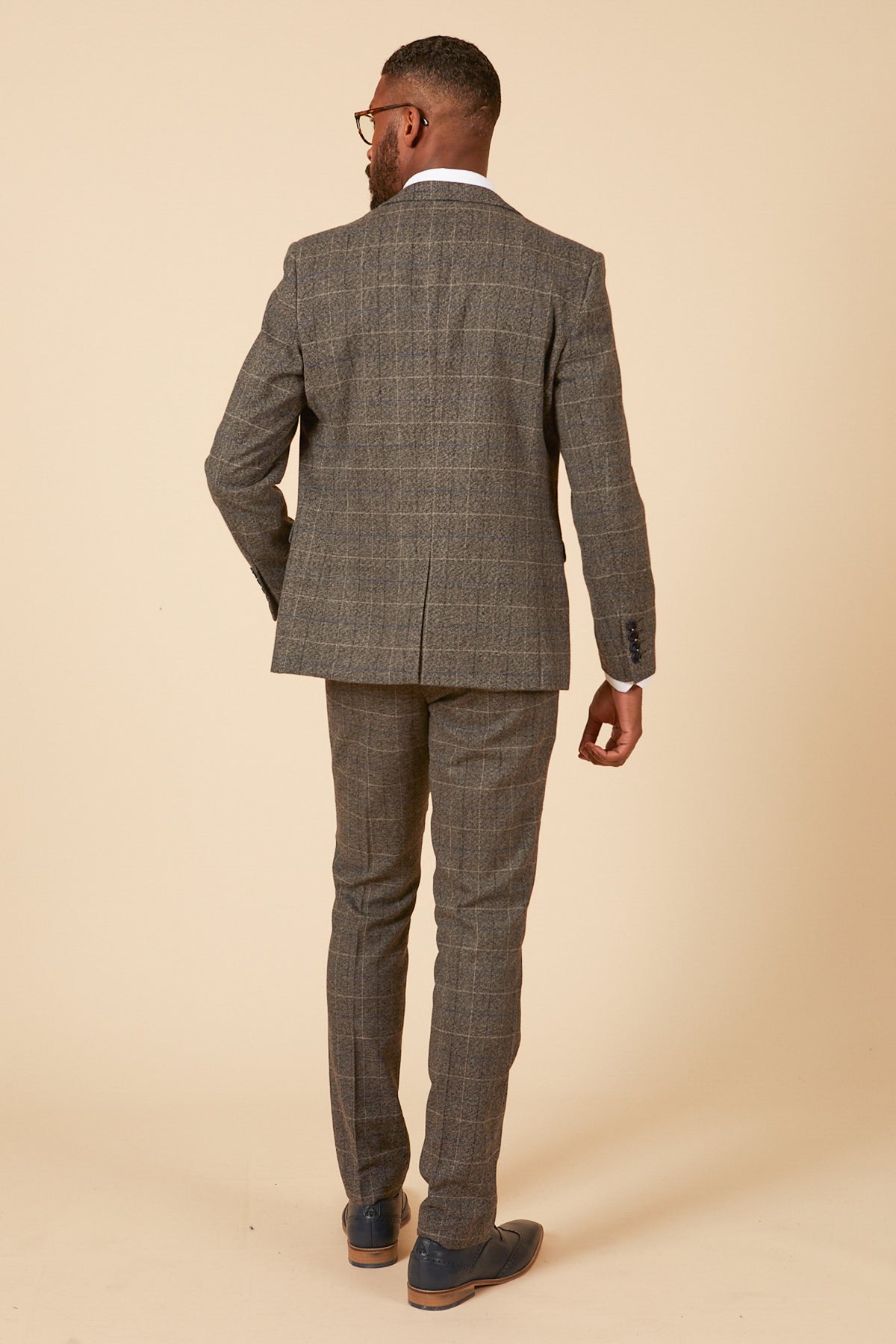 A man with short hair, glasses, and a beard models the marcdarcy SCOTT Grey Tweed Check Three Piece Suit with navy velvet pocket trims, white shirt, and dark loafers against a plain beige background.