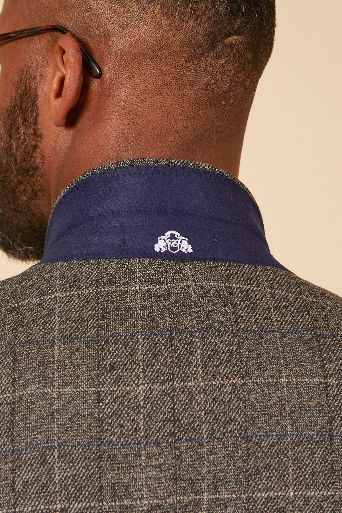 A close-up of the back of a mans neck wearing marcdarcys SCOTT Grey Tweed Check Blazer, featuring a blue collar with navy velvet trim and a small white embroidered crest for a vintage-inspired look.
