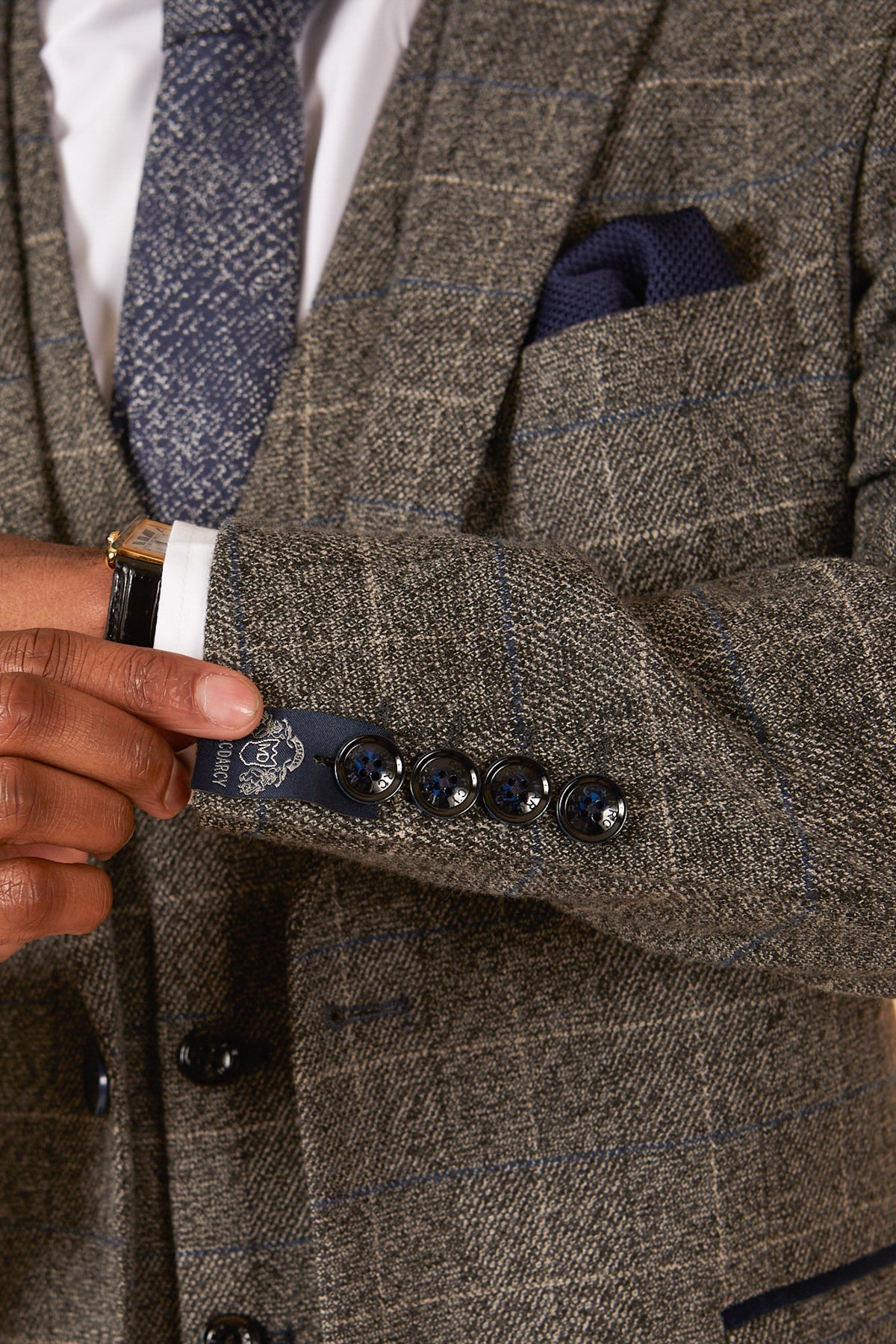 A close-up of a hand adjusting the cuff of a marcdarcy SCOTT Grey Tweed Check Blazer with four dark buttons, paired with a white shirt, patterned tie, and matching navy blue pocket square.