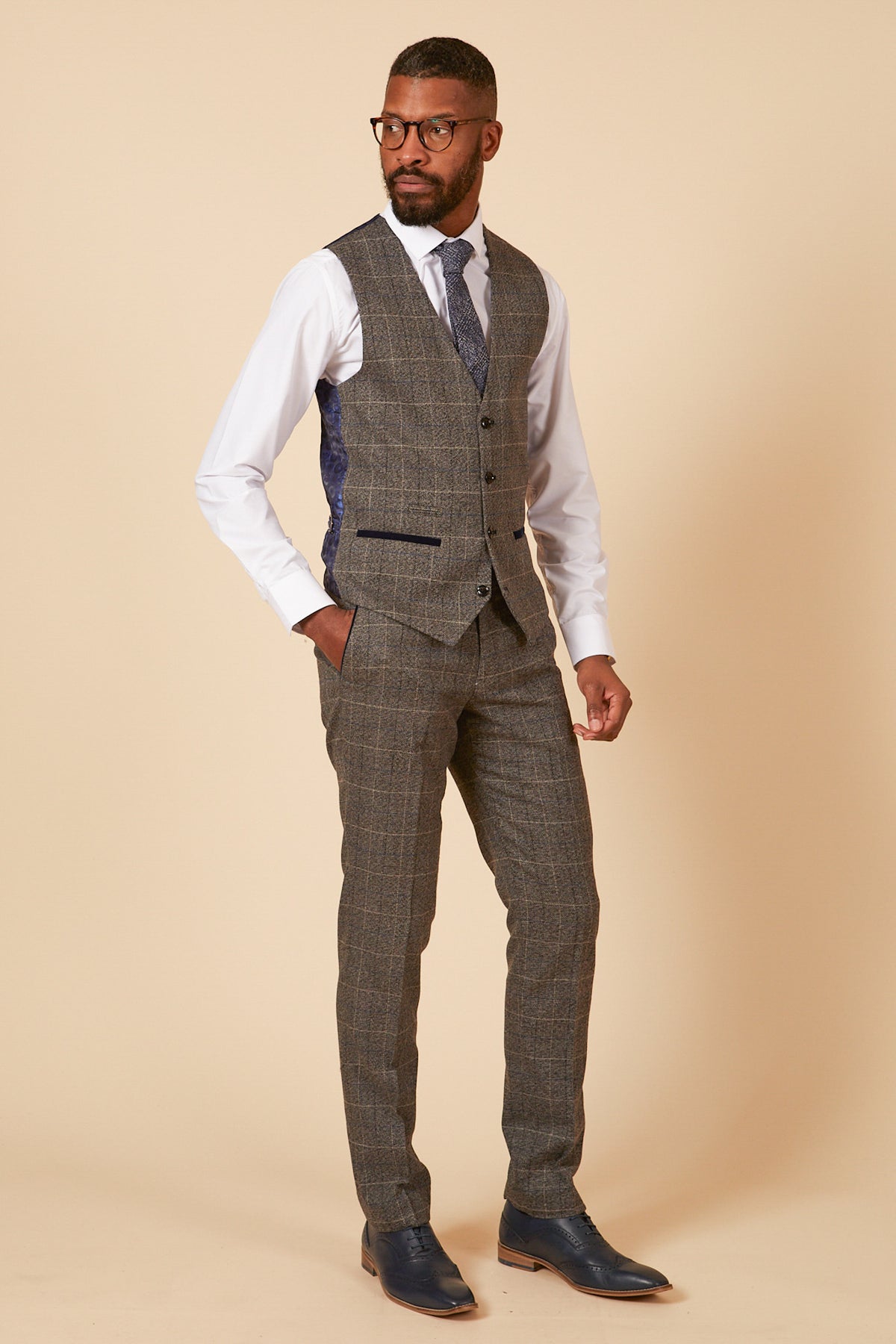 A man wearing glasses, a white dress shirt, tie, and the marcdarcy SCOTT - Grey Tweed Check Three Piece Suit stands against a plain beige background.