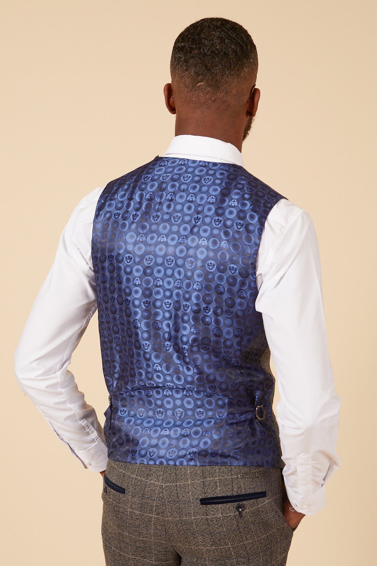 A man facing away wears the marcdarcy SCOTT Grey Tweed Check Three Piece Suit, featuring a white dress shirt, grey plaid trousers, and a shiny blue vest with circular patterns and navy velvet pocket trims. Plain beige background.