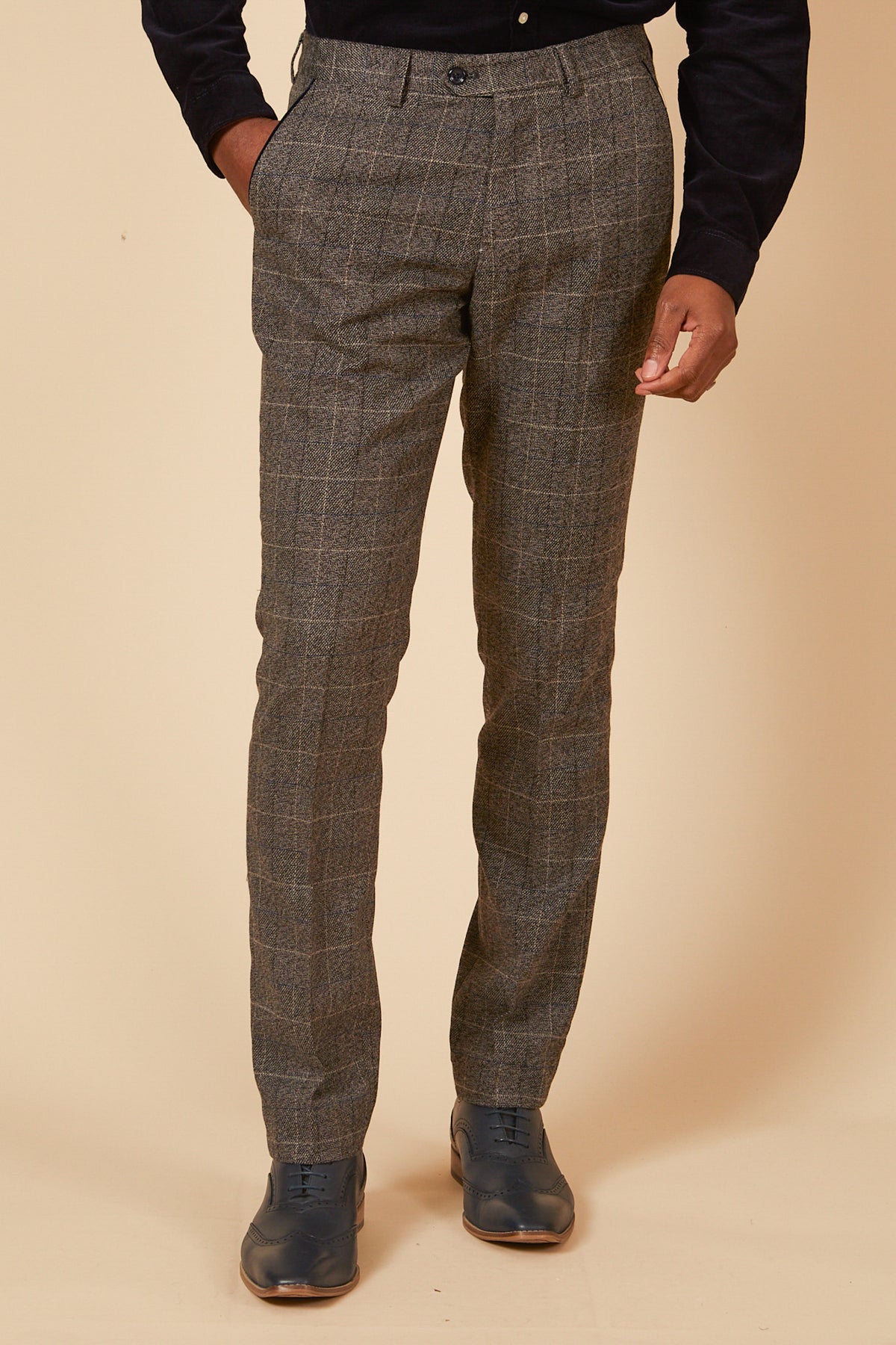 A person stands against a beige background wearing marcdarcy SCOTT Grey Check Tweed Trousers, a dark long-sleeve shirt, and dark shoes, with one hand in their pocket.