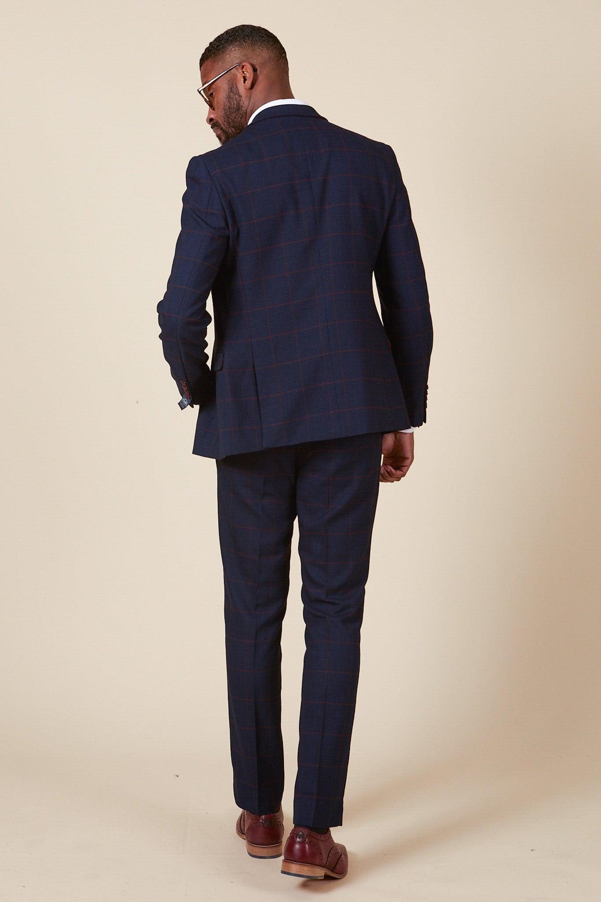 A man in the EDINSON - Navy Wine Check Two Piece Suit by Marc Darcy stands with his back to the camera, head turned left. He completes the look with brown shoes and glasses against a plain beige background.