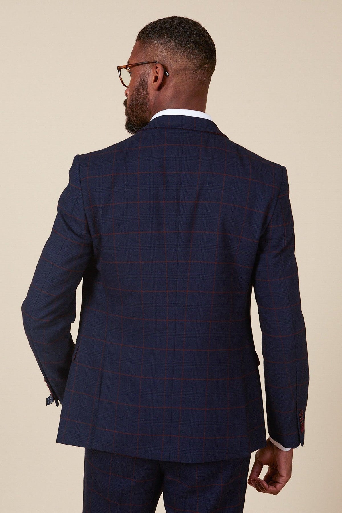 A man with short hair, a beard, and glasses is seen from behind wearing the Marc Darcy EDINSON Navy Wine Check Two Piece Suit, featuring a subtle red check pattern. The plain background highlights his stylish look.