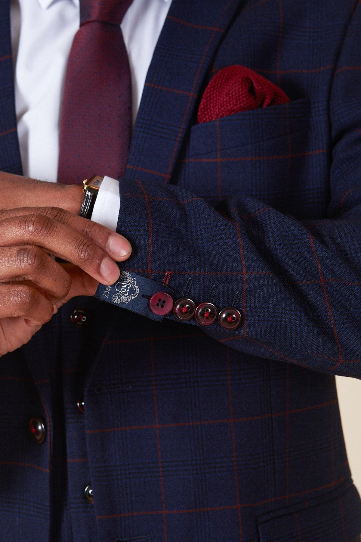 A person in the Marc Darcy EDINSON Navy Wine Check Two Piece Suit adjusts their sleeve, showcasing a white shirt, burgundy tie and pocket square, gold watch, and a slim fit blazer with four buttons.